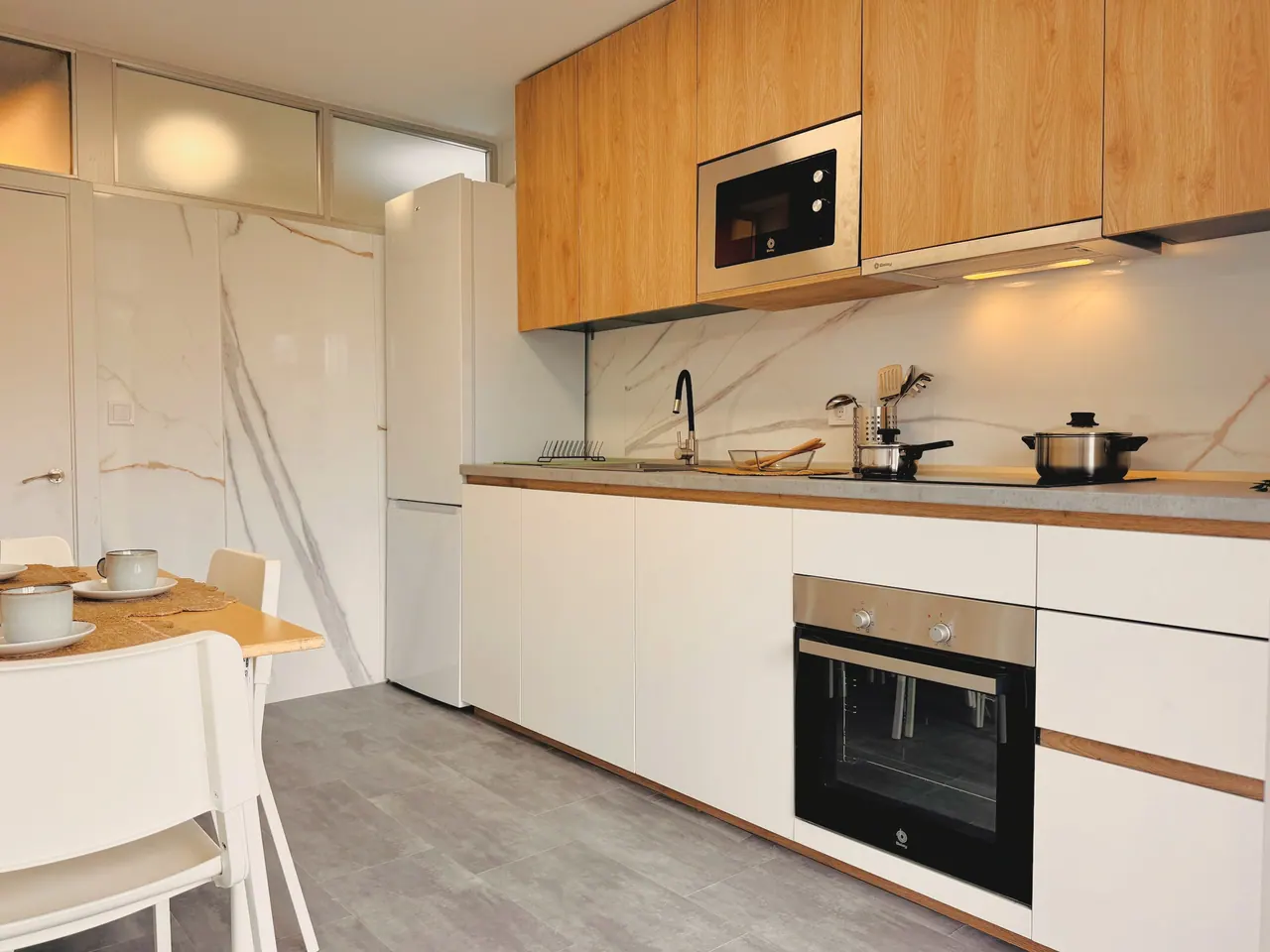 Cocina con muebles de madera y blancos, horno, microondas, frigorífico y mesa con sillas blancas