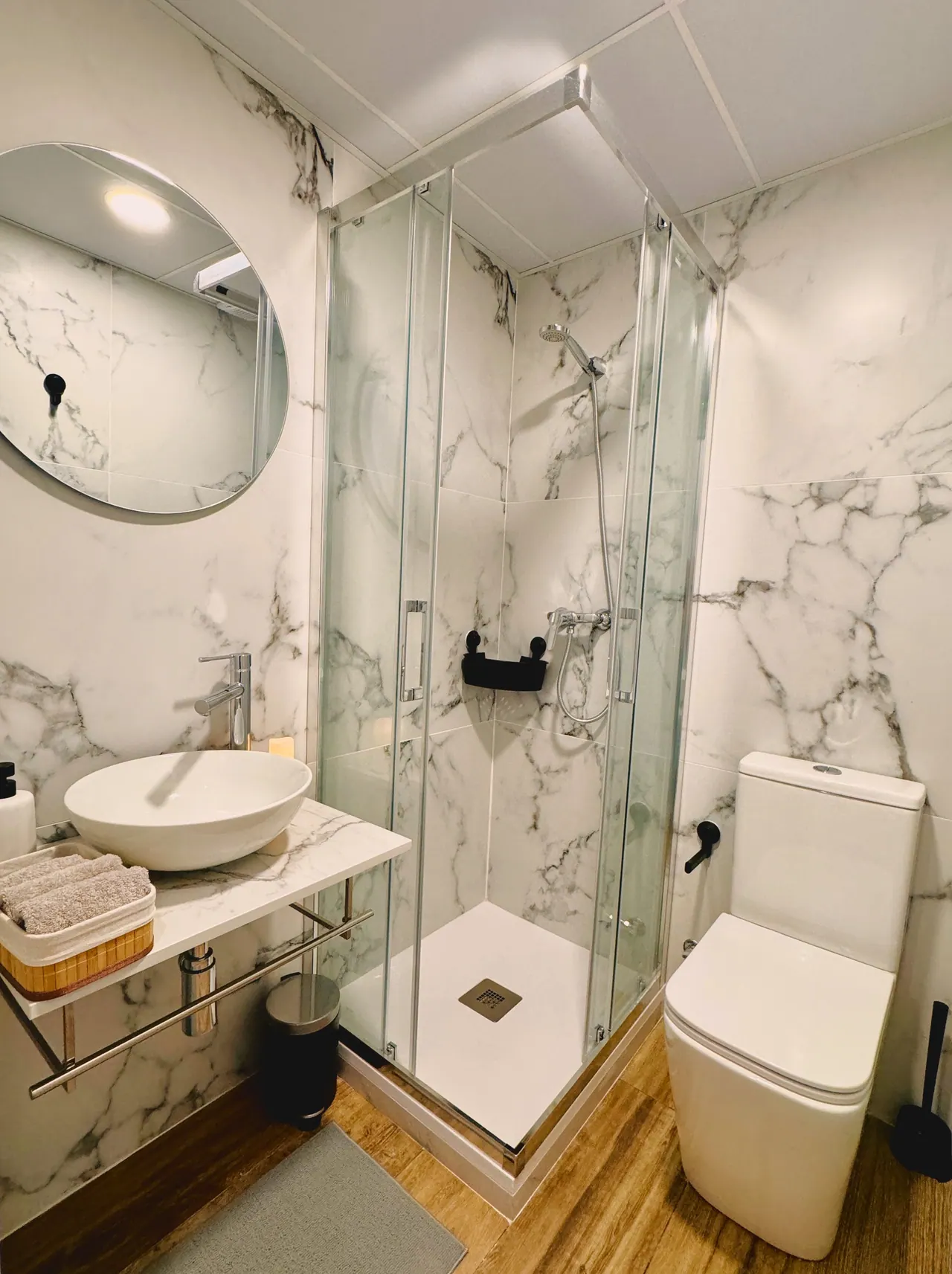 Baño de mármol blanco con lavabo sobre encimera, espejo redondo y ducha con mampara (foto 3 de 8)
