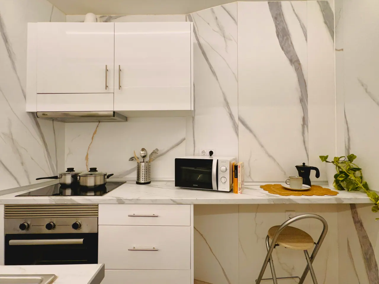 Cocina blanca con pared de mármol, placa, horno, microondas y taburete plegable