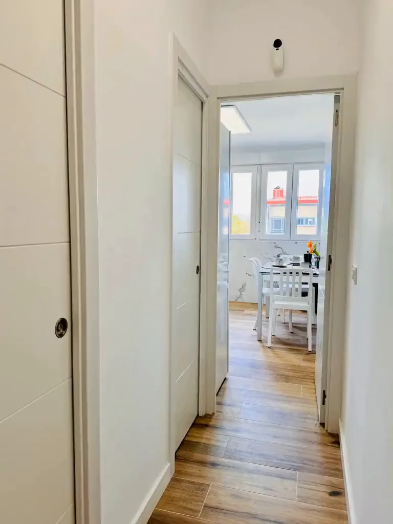 Pasillo blanco con suelo de madera que lleva a una cocina luminosa con mesa y ventanal